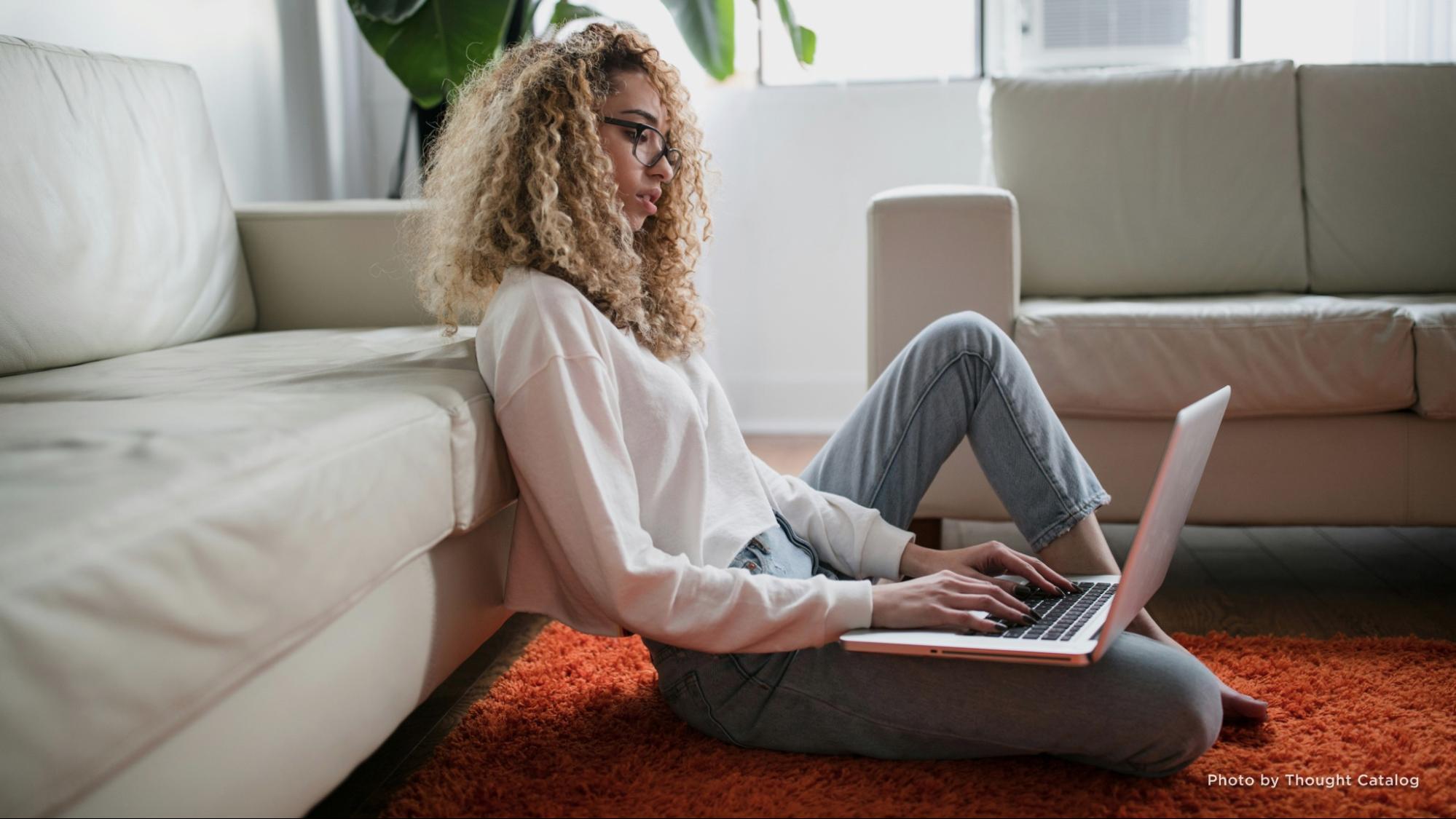 Woman sitting on the floor and leaning against a couch while using a laptop.