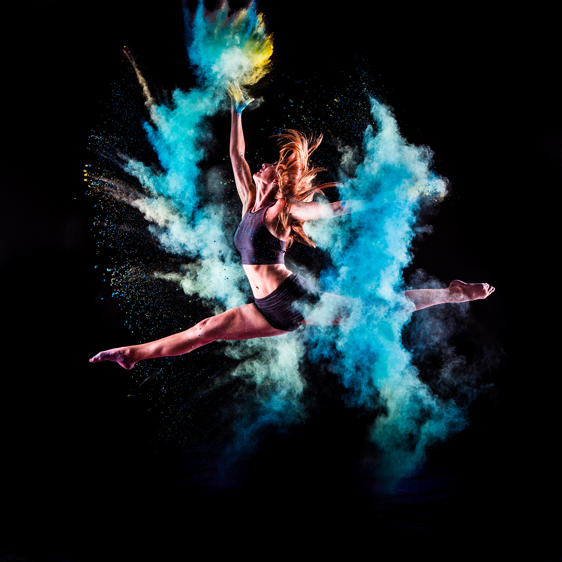 Dancer leaping in the air against a black background and a colorful spray of blue powder.